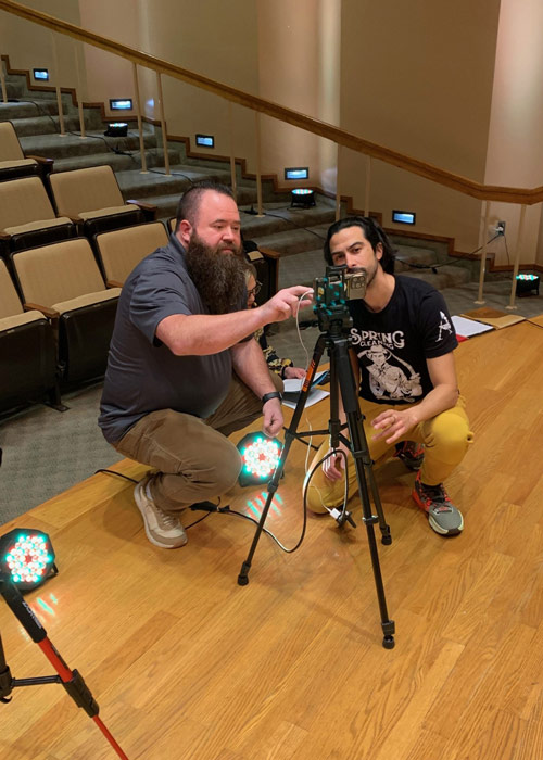 Producer Lenny Hernandez and Videographer Colby Boyes kneel on a wooden stage, adjusting cameras and lights before filming Thoughtful Connections: A Memory Care Podcast.