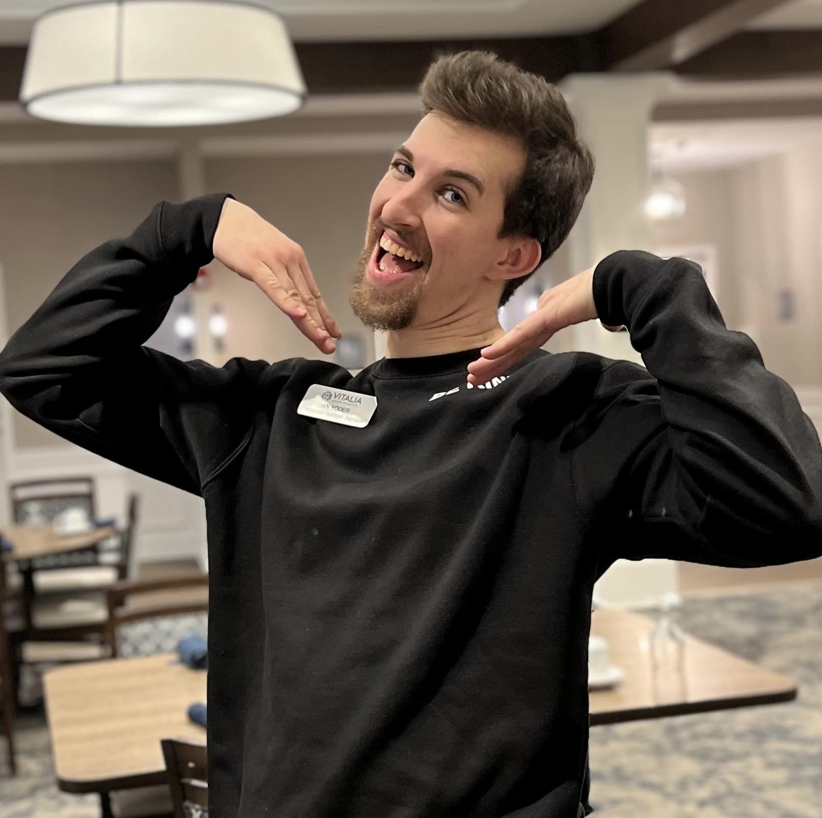Senior living team member playfully poses with a bright smile, hands under chin, while wearing a name tag and black sweater, showcasing enthusiasm and joy in the community setting.