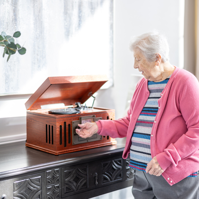 A senior resident in a pink cardigan operates a vintage record player on a decorative table, enjoying a nostalgic moment in a well-lit room with soft natural light.