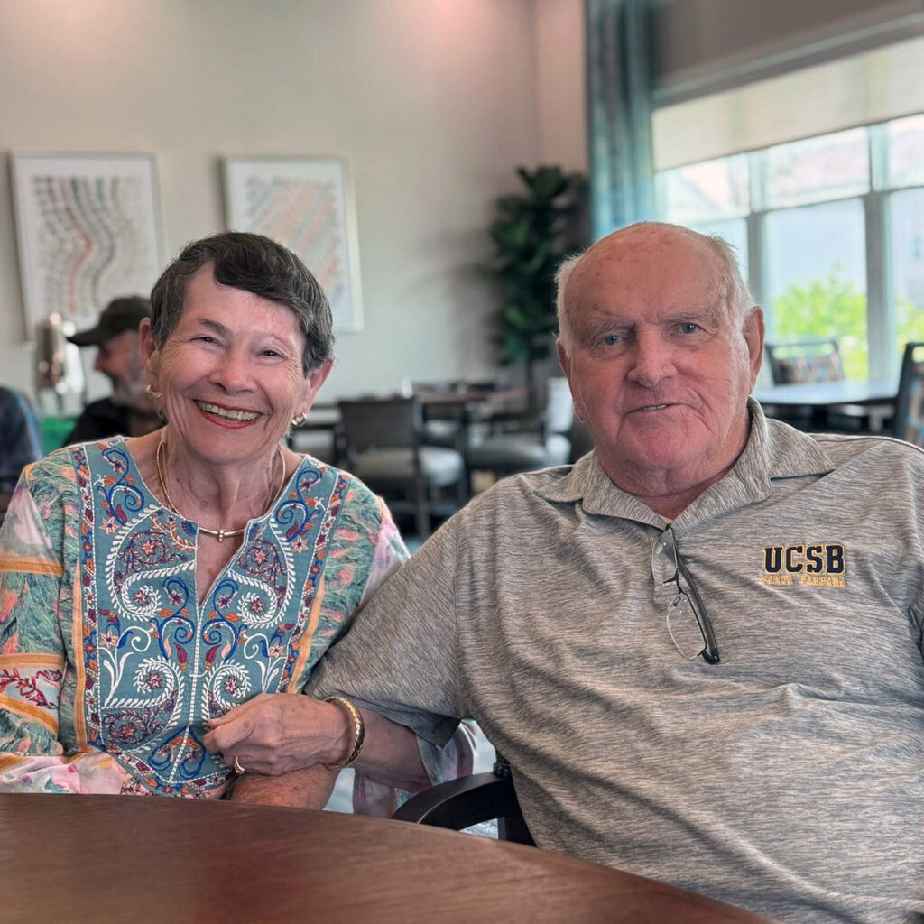 Senior woman in a colorful blouse smiles seated at a table next to a senior man in a gray shirt.