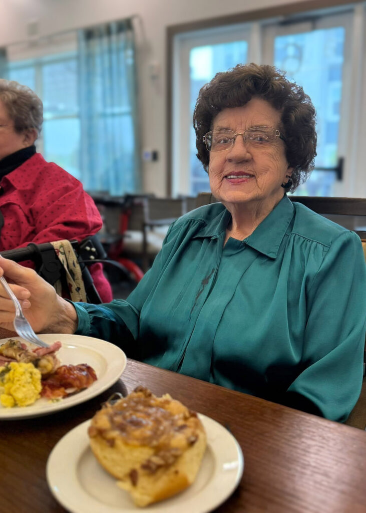A senior resident wearing a teal blouse smiles while enjoying a meal of eggs, bacon, and a cinnamon roll at a dining table in the community.
