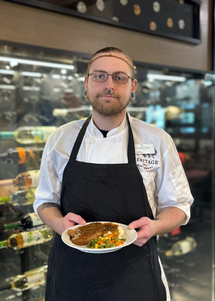 A Culinary Director in a chef coat holds a plate of meatloaf with mashed potatoes and vegetables, standing in front of a wine rack display in the dining area.