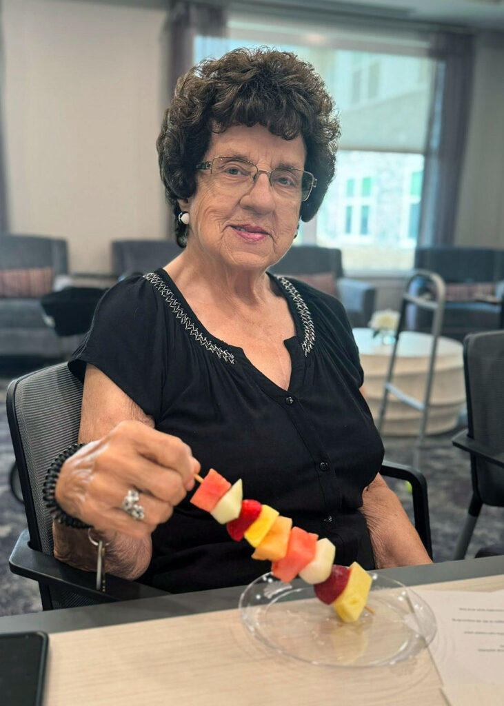 A senior woman smiles at a dining table, holding a skewer of assorted fruits.