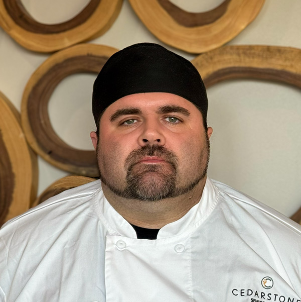 David Klimesh, Culinary Director at CedarStone Senior Living, in a professional headshot. He has short dark hair under a black chef hat, a trimmed beard and mustache, and wears a white chef coat with the CedarStone logo, standing in front of a modern wood-patterned background.