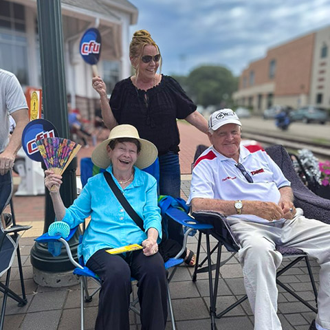 Team member smiles with two senior residents seated in lawn chairs during an event outdoors.