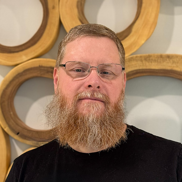 Joe Elliott, Plant Operations Director at CedarStone Senior Living, pictured in a professional headshot. He wears glasses and a black shirt, with a decorative wood circle wall background.